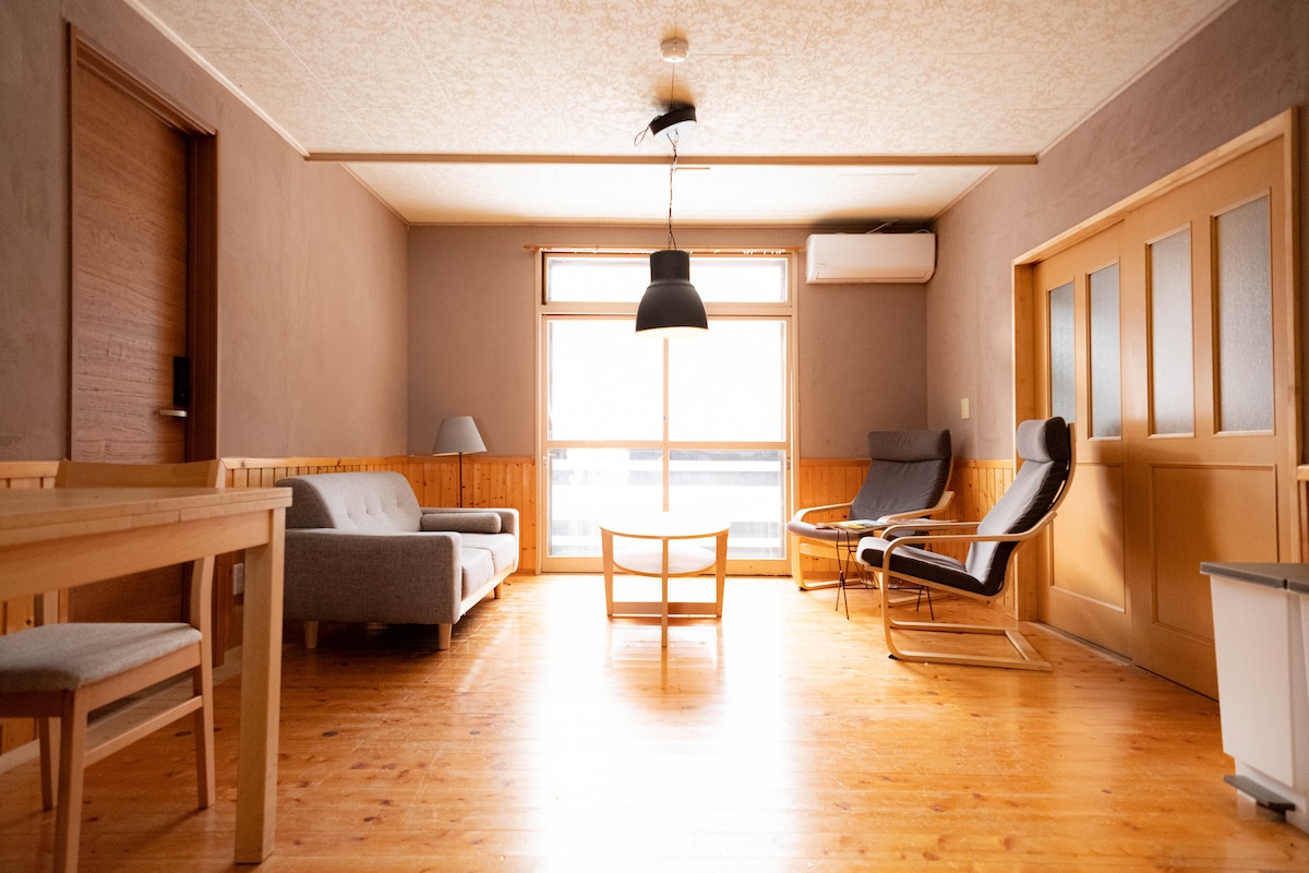 A spacious lounge area features natural wood flooring and soft lighting. A large window allows ample sunlight, illuminating a minimalist seating arrangement that includes a gray sofa and two comfortable armchairs. A simple wooden table is placed centrally, enhancing the room's inviting atmosphere.