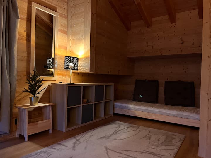 Esquina de montaña abierta (relax), por la noche, con la 2ª cama individual-banqueta y sus 2 cojines cuadrados. Luz suave tamizada para crear un ambiente relajante.