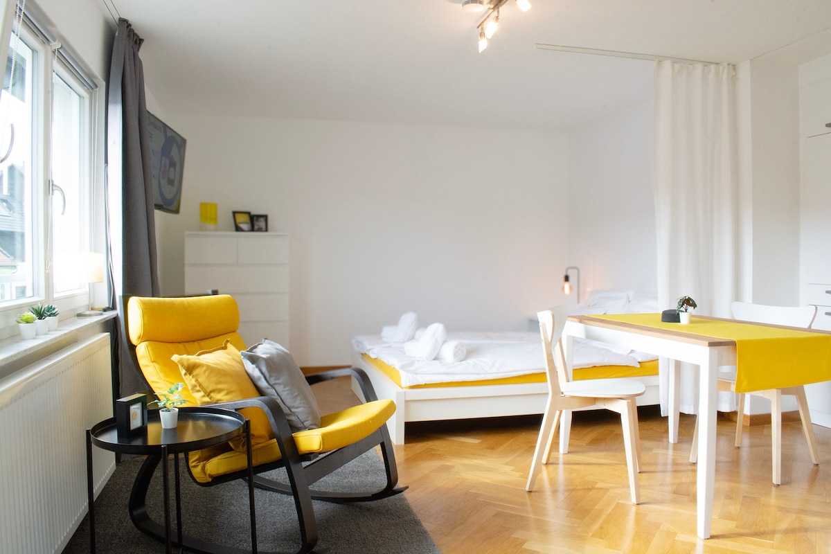 A bright living space features a modern yellow lounge chair beside a small black table. A dining area with a white table and chairs complements the room. In the background, a spacious bed is neatly made, with soft linens inviting rest.