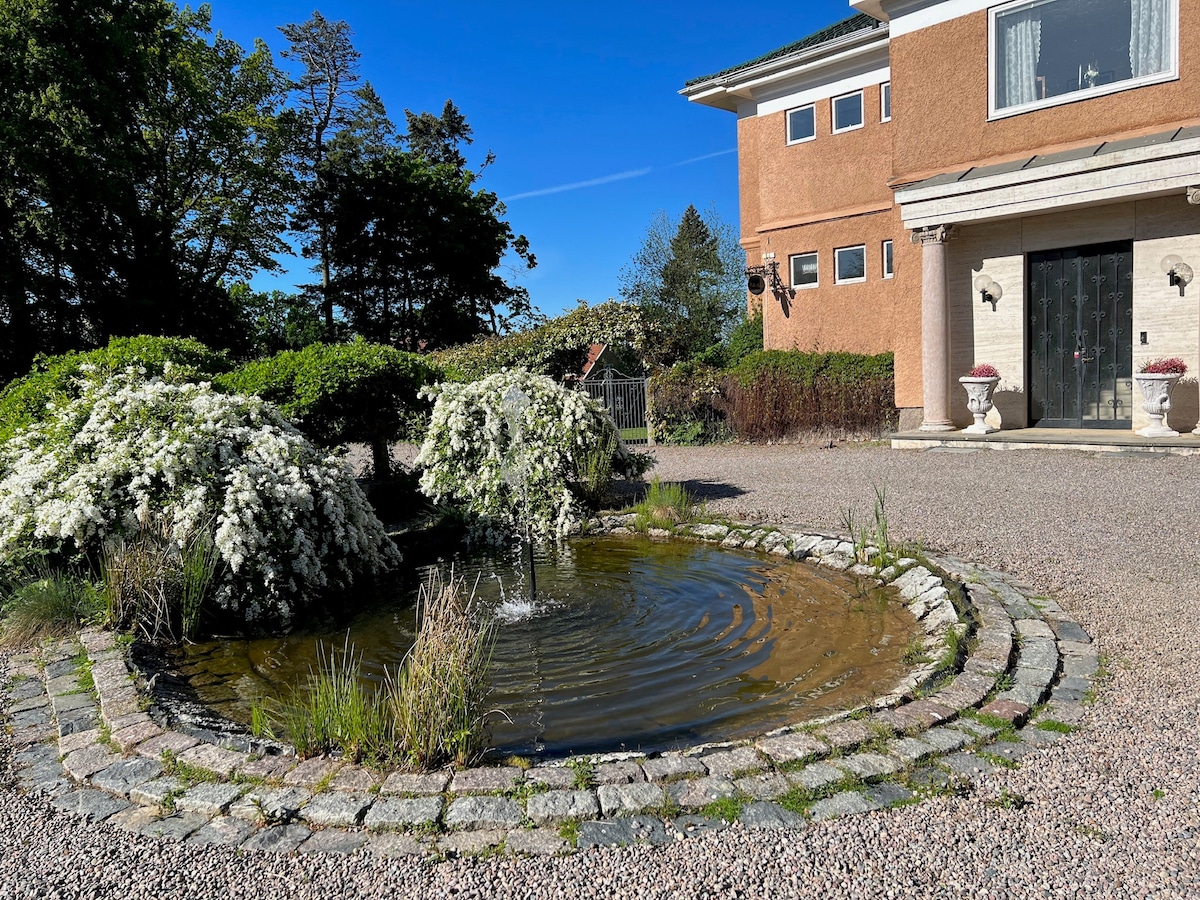 A serene garden area features a circular stone fountain surrounded by lush greenery and flowering plants. The building's entrance is visible in the background, highlighting the spacious layout. A clear blue sky enhances the tranquil outdoor environment.