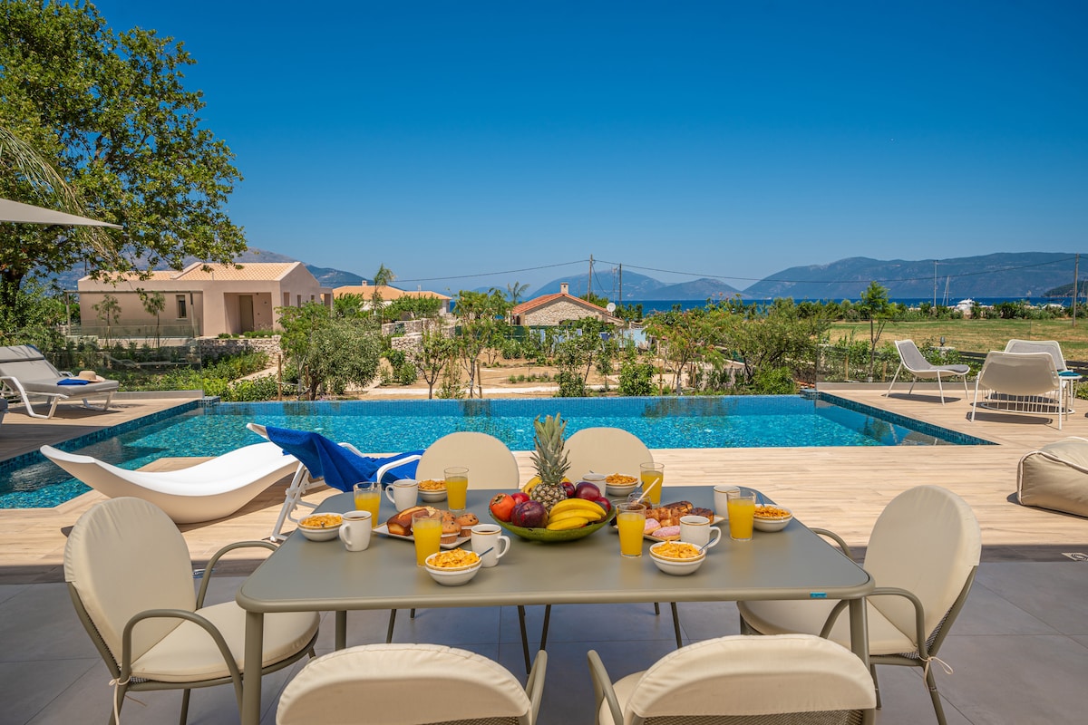 An outdoor dining area features a table set with various breakfast items, including fresh fruit and drinks. The private pool glimmers nearby, surrounded by comfortable loungers, with views of the sea and mountains in the distance under a clear blue sky.