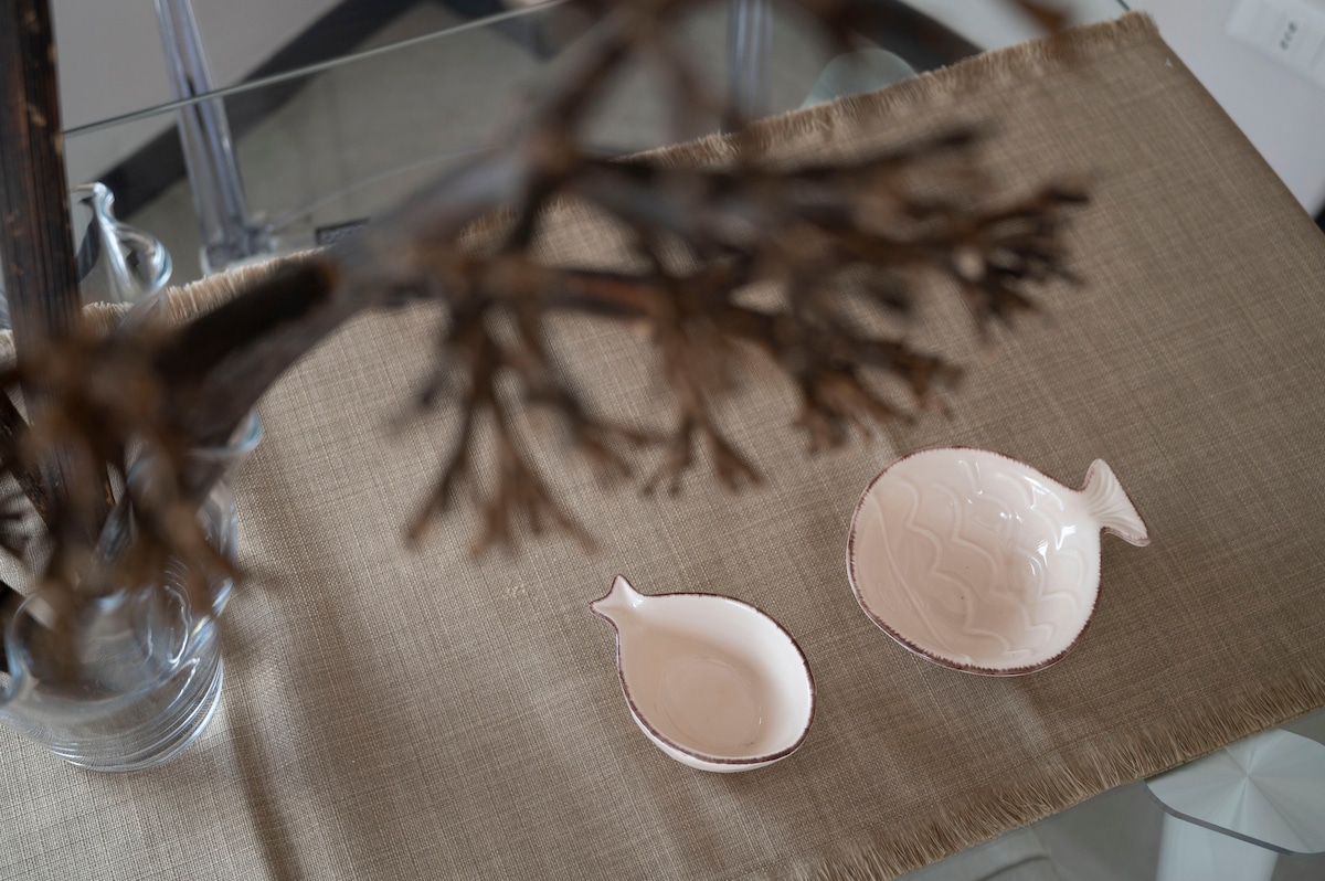 A contemporary dining table is adorned with a light-colored table runner. Two ceramic fish-shaped dishes in soft pink tones are positioned on the runner, surrounded by a minimalist floral arrangement that adds a touch of nature to the setting.
