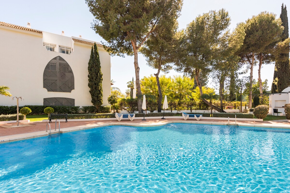 A large outdoor swimming pool is surrounded by well-maintained gardens and tall trees. Several sun loungers are positioned near the water, offering a relaxing atmosphere. The building is visible in the background, enhancing the tranquil outdoor setting.