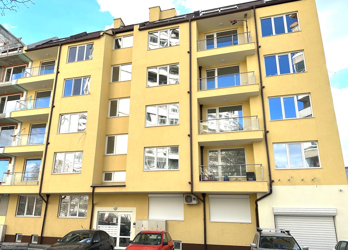 A modern, newly constructed apartment building is shown in a light yellow exterior. Large windows allow ample natural light, and several balconies are visible. The entrance features double doors, with a few cars parked in front, demonstrating convenient access and proximity to free parking.