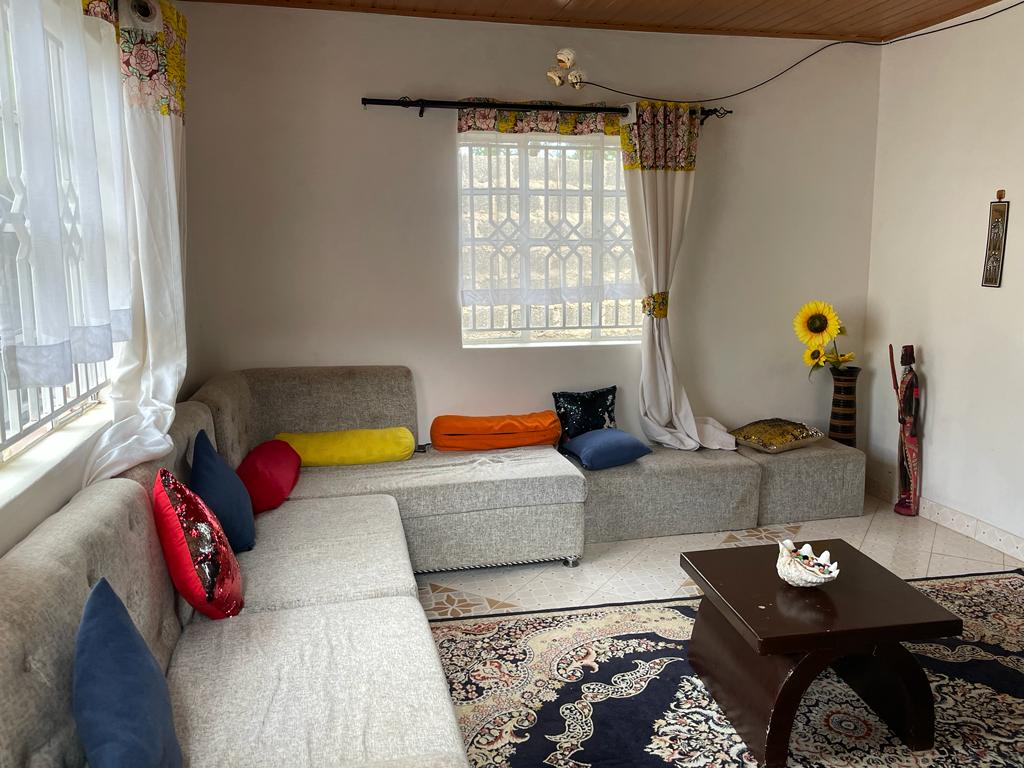 A spacious living area features a light grey sectional sofa adorned with colorful cushions. A low dark wood coffee table is placed on a patterned area rug. Large windows provide natural light, complemented by bright, floral drapes and a vibrant sunflower decor.