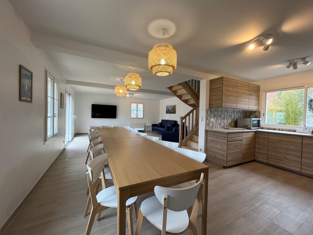 A spacious dining area features a long wooden table surrounded by multiple white chairs, designed for group gatherings. Soft pendant lights hang from the ceiling, providing an ambient glow. The open layout connects seamlessly with the kitchen, showcasing modern cabinetry and a cozy seating area.