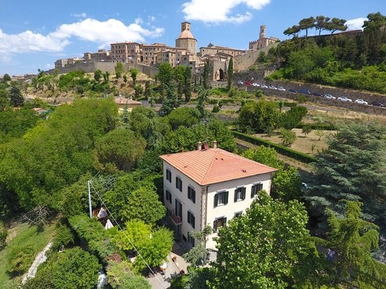 A charming villa is situated amidst lush greenery, with a view of the historic town in the background. The structure features a classic façade, red tiled roof, and numerous trees framing the property, creating a serene environment close to the ancient city walls.