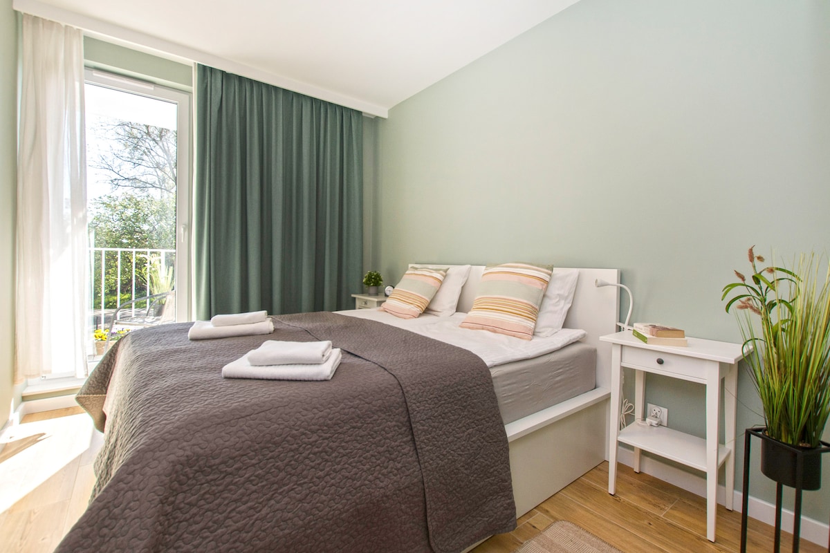 A spacious sleeping area features a double bed with gray bedding and decorative pillows. Natural light floods in through a glass door leading to a balcony, with green curtains partially drawn. A small bedside table and a potted plant enhance the space.