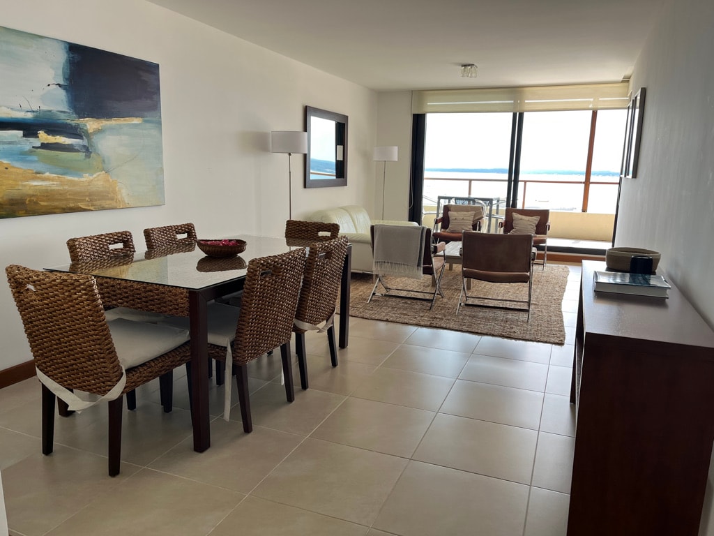 A spacious dining area is furnished with a rectangular table surrounded by six woven chairs. The living area features a light-colored sofa and two armchairs, all situated on a neutral-toned area rug. Large windows offer views of the bay, enhancing the bright and airy ambiance.