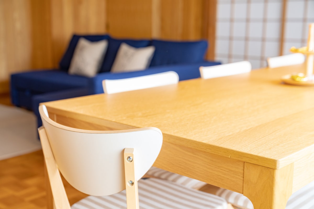 A dining area features a wooden table with a light finish and white chairs. In the background, a blue sofa with two decorative pillows is visible, adding to the inviting atmosphere of the space. Natural light enhances the warm tones of the wood.