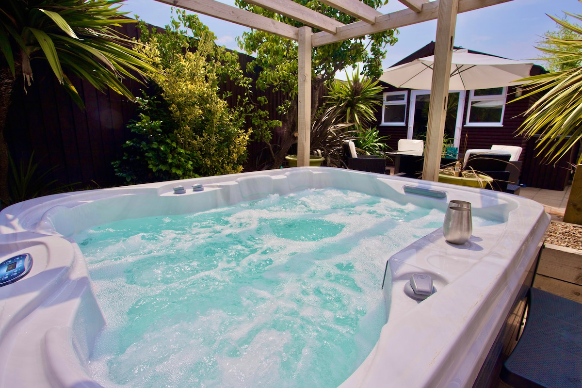 A luxurious hot tub is positioned under a wooden pergola, surrounded by lush greenery and tropical plants. The bubbling water reflects sunlight, creating a tranquil atmosphere. Comfortable outdoor seating is visible in the background, offering a serene space for relaxation.