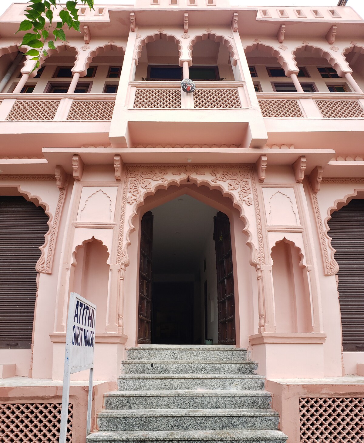 The entrance of a guest house features intricate pink architectural details and arched doorways. Stone steps lead up to the grand entrance, which is framed by wooden doors. A sign indicating the guest house is placed at the foot of the stairs.