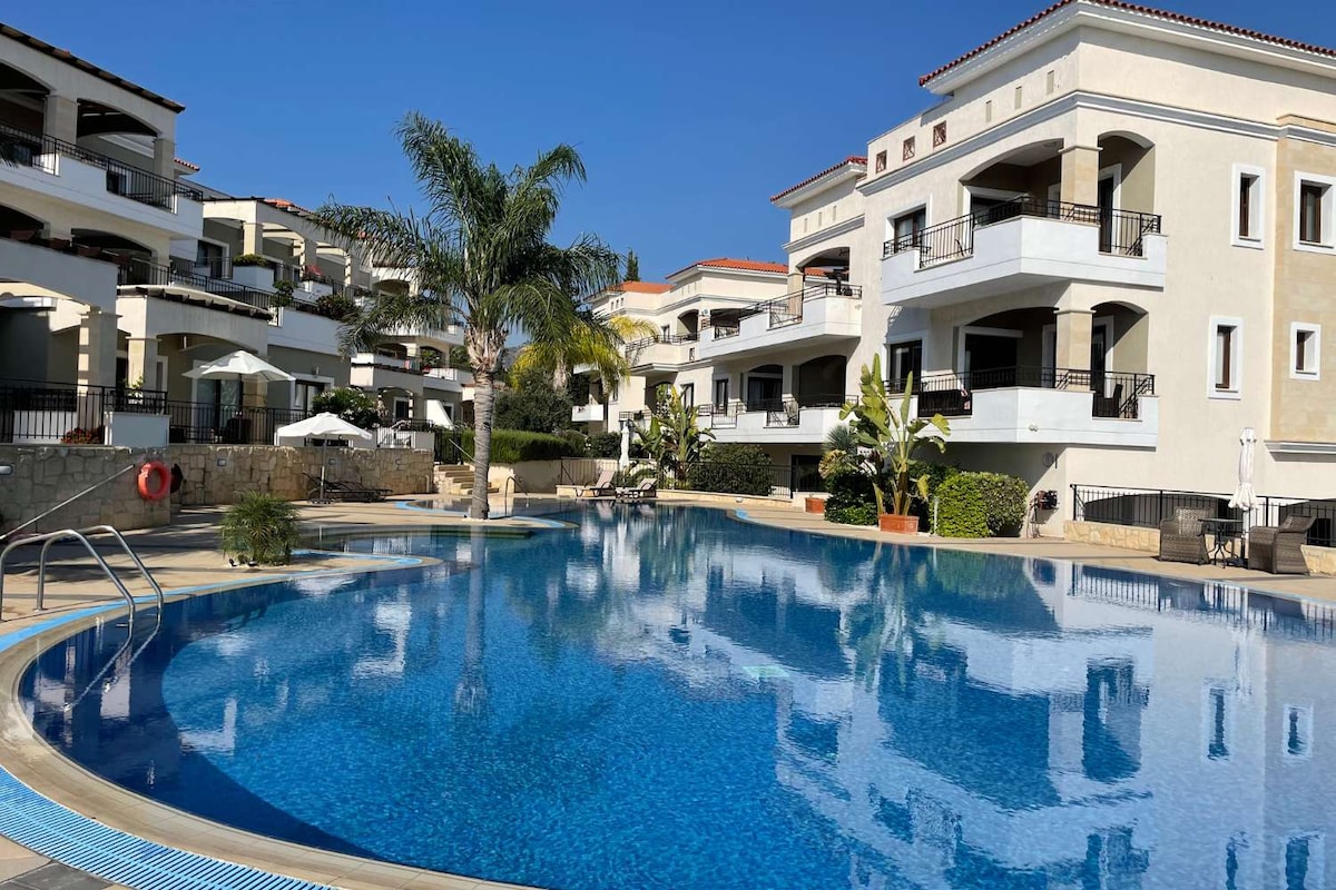 The image depicts an outdoor infinity pool surrounded by lush greenery and modern apartment buildings. Palm trees frame the scene, and the pool's water reflects the clear blue sky. Comfortable sun loungers are positioned nearby, contributing to a relaxing atmosphere.