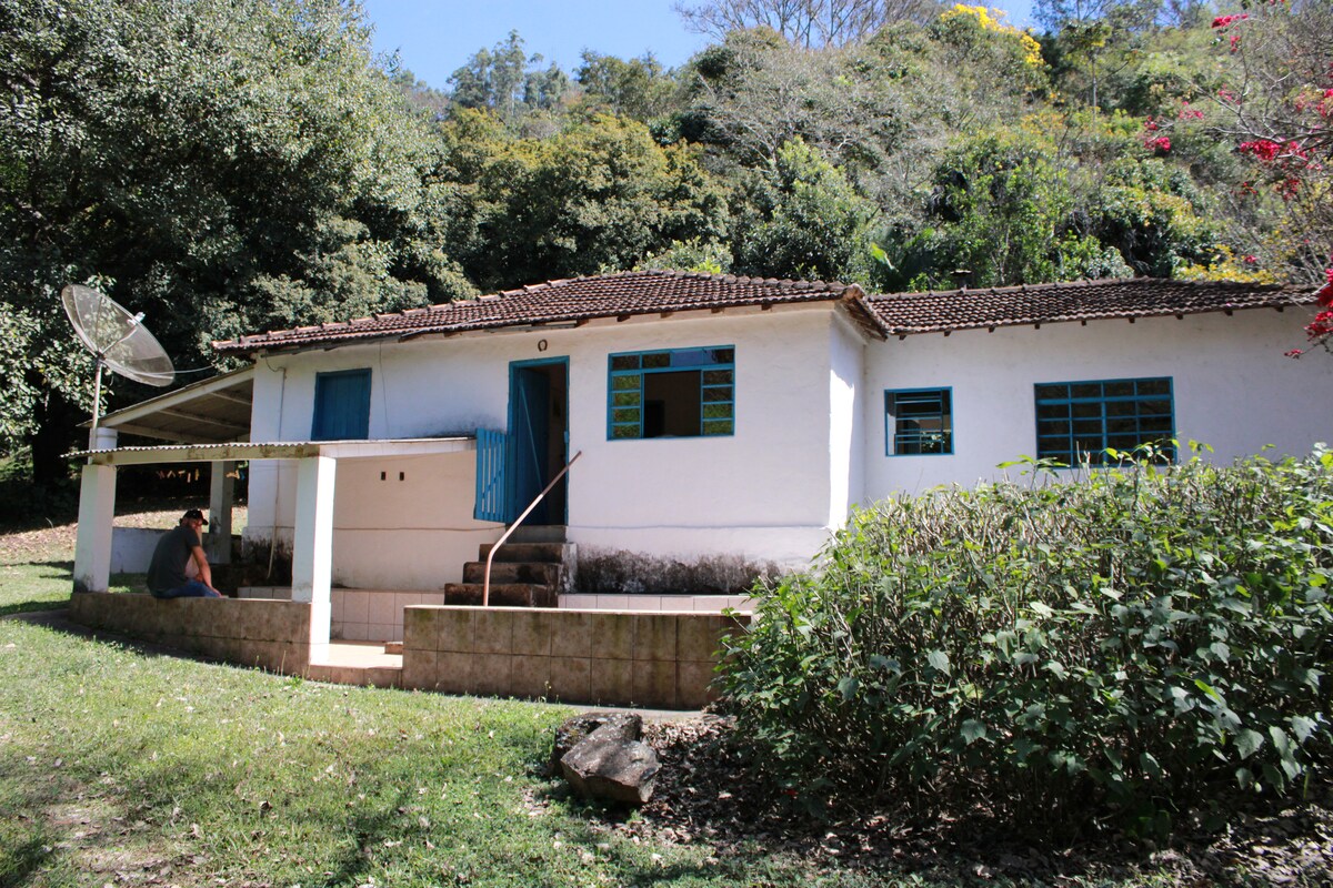 A single-story house is situated amidst lush greenery. The exterior features a whitewashed facade with blue windows and a welcoming porch. Stone steps lead to the entrance, while the surrounding garden is filled with various plants and trees, enhancing the natural setting.
