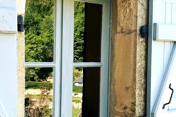 A window frame is seen from the outside, featuring a light blue color with wooden shutters. The glass reflects the lush greenery of the surrounding landscape, inviting a sense of tranquility and connection to nature.