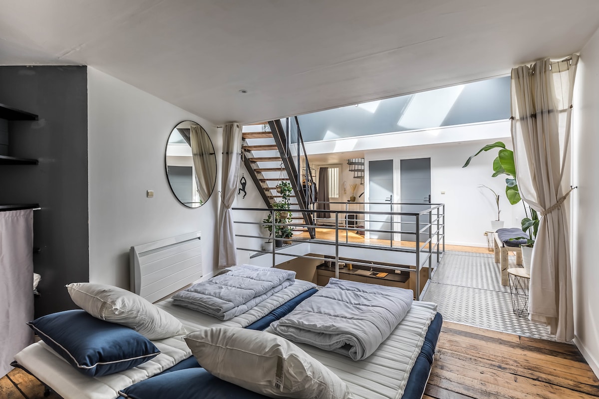 A spacious sleeping area features a large bed with fresh bedding and a circular mirror on the wall. Light enters through a skylight above, illuminating the open space and a staircase leading to the upper level. A plant adds a touch of greenery.