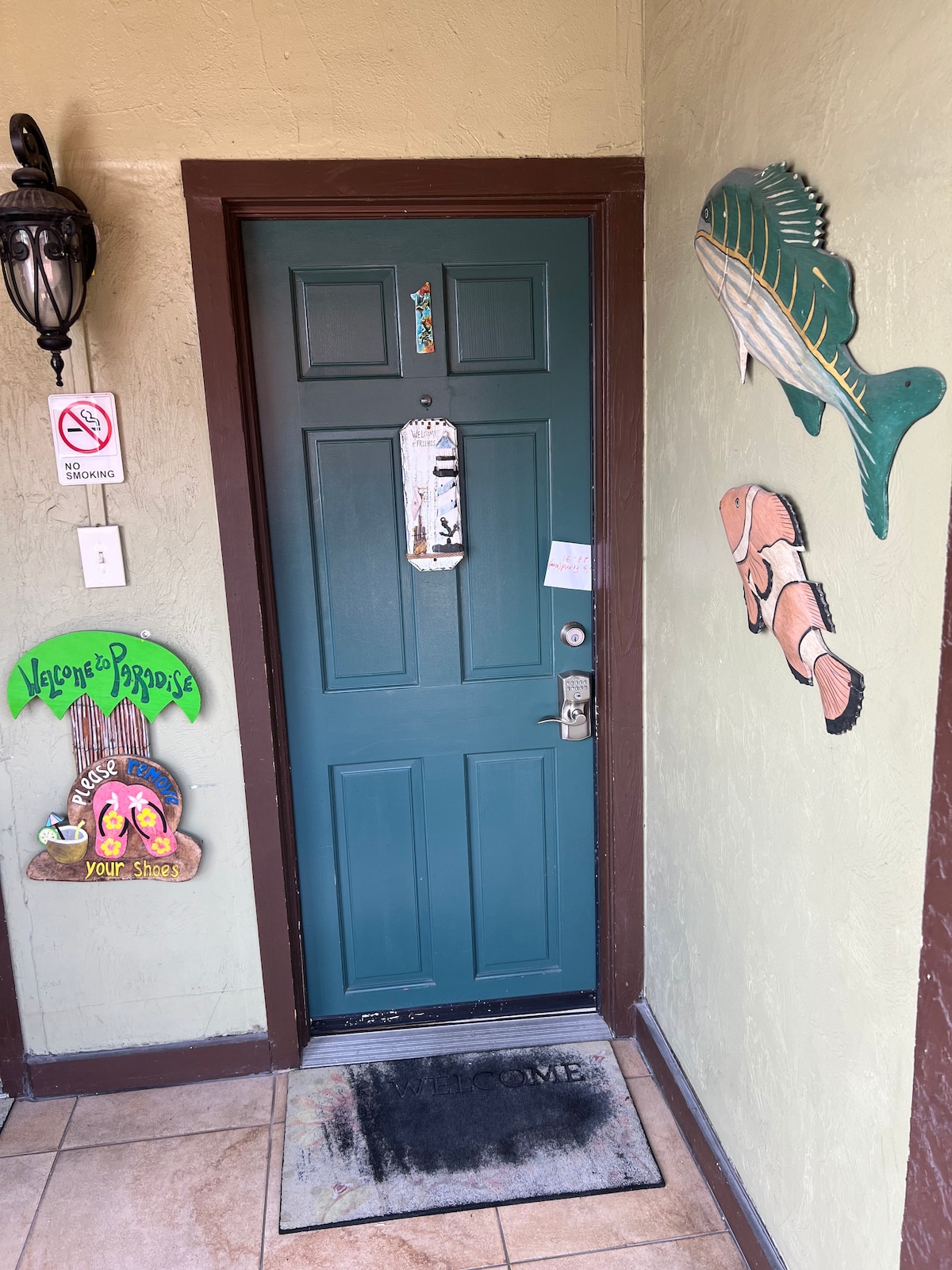 A vibrant entry door is framed by painted wall decor, including marine-themed signs. A doormat reading 'WELCOME' is positioned at the entrance, inviting guests into the space. Soft lighting highlights the textured wall surface and decorative elements around the door.