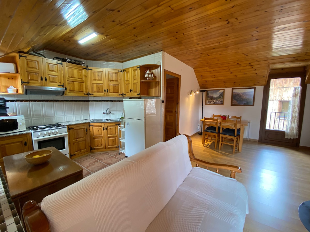 An open kitchen area with wooden cabinetry is visible alongside an adjacent dining space. A light-colored sofa is positioned in the foreground, while a round dining table with chairs can be seen in the background, illuminated through a window with wooden shutters.