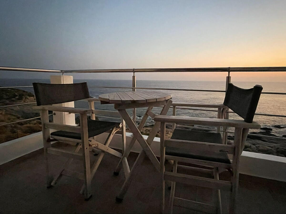 A small outdoor balcony features two wooden chairs and a round table, positioned to overlook the sea. The early evening sky displays soft hues as the sun sets, creating a serene backdrop for relaxation.