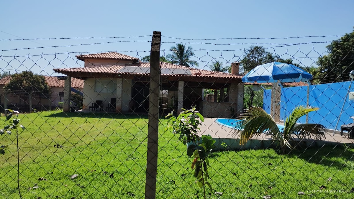 The exterior view showcases a single-story house with a tiled roof and a fenced yard. A well-maintained lawn surrounds the property, featuring a pool area with a blue cover and a shaded seating space under an umbrella. Palm trees are visible in the background.