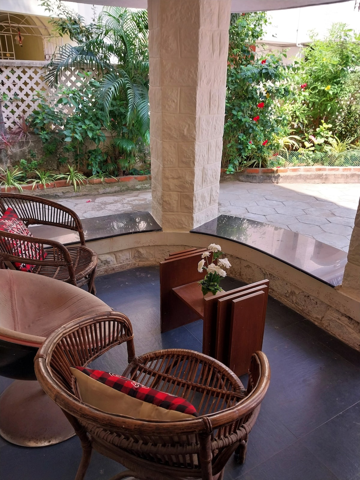 A seating area is surrounded by lush greenery, featuring two woven chairs and a small wooden table. A decorative plant sits atop the table, with red flowers adding a pop of color in the background. The space is well-lit, creating an inviting and serene environment.