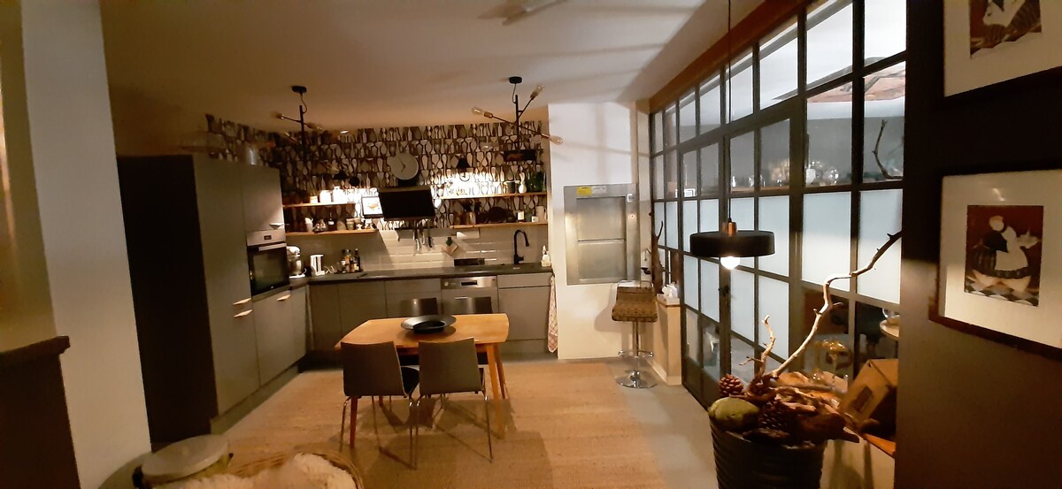 A modern kitchen is depicted with sleek cabinets and stainless steel appliances. A wooden dining table with two chairs is positioned in the center, complemented by a textured rug. Large windows allow natural light to fill the space, enhancing the warm, inviting ambiance.