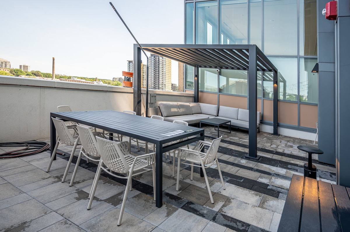 A spacious rooftop terrace features a modern pergola providing shade over a seating area. A rectangular table with eight surrounding chairs is set on a stone-paved surface, offering ample space for outdoor gatherings while enjoying the city views.