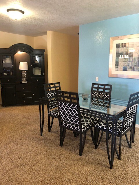 A dining area is displayed, featuring a glass-top table surrounded by six black chairs with a geometric pattern. A dark cabinet is positioned against the wall, complemented by a lamp and framed artwork, enhancing the space's contemporary design.