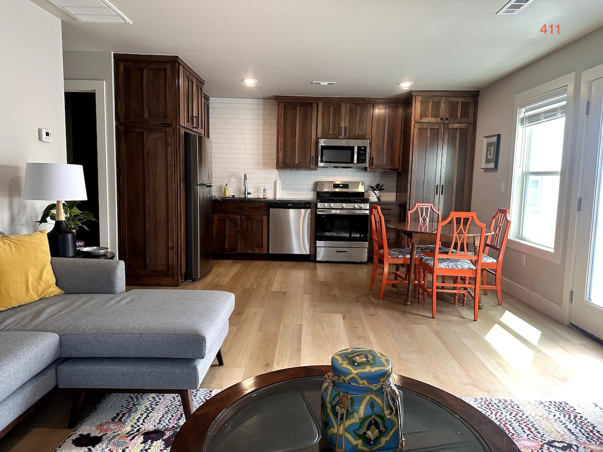 An open living space features a modern kitchen with stainless steel appliances and wooden cabinetry. A dining area with four chairs is positioned next to the kitchen. A comfortable gray sofa is placed in the foreground, complemented by a decorative table with an eye-catching centerpiece.