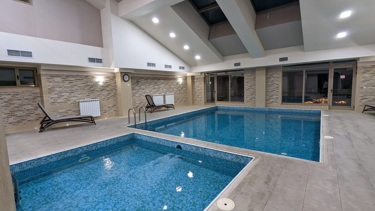 An indoor swimming area is shown, featuring two pools with clear blue water. Surrounding the pools are lounge chairs arranged for relaxation. The room is well-lit, with high ceilings and large windows providing natural light. Wall finishes include stone and tile elements.
