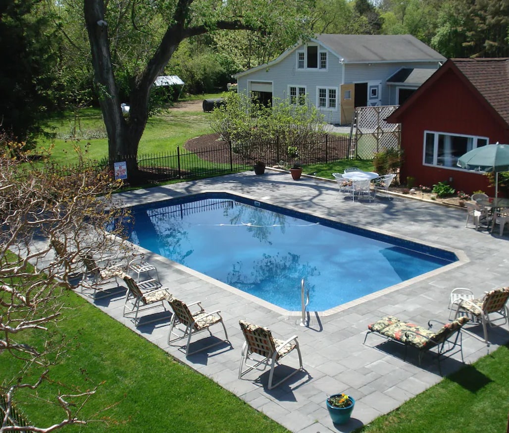 An outdoor pool area is featured, surrounded by a paved deck and lounge chairs. A bright blue pool is the focal point, with greenery and trees visible in the background. Nearby tables and an umbrella offer shaded seating options.