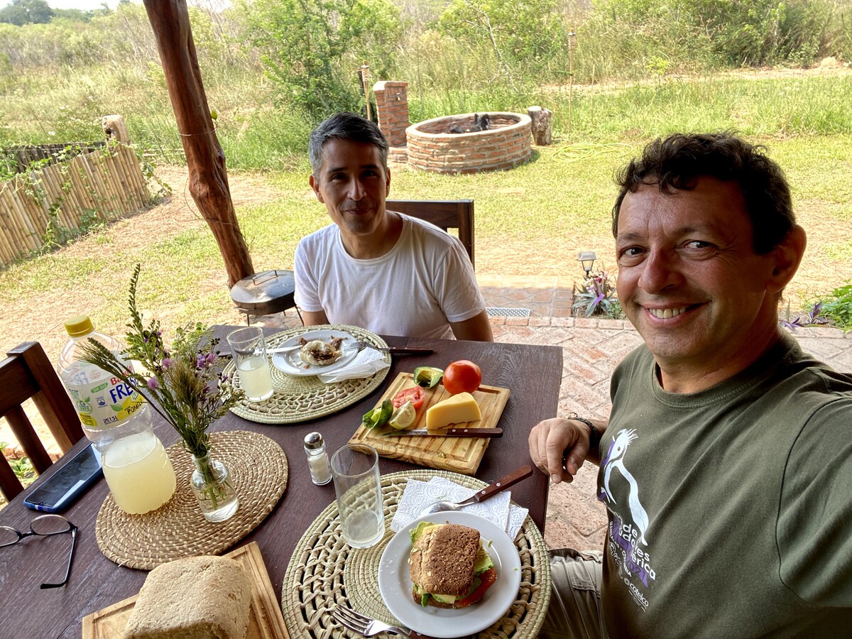 A table is set outdoors with two plates featuring sandwiches, cheese, and fresh fruits. Glasses of lemonade are placed alongside, while a scenic view of greenery is visible in the background. A stone fire pit can be seen further back, enhancing the natural setting.
