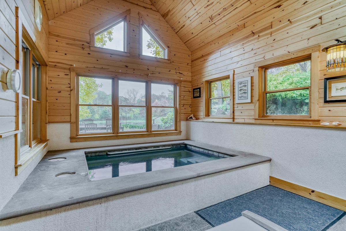 An indoor spa area features a small jacuzzi, surrounded by wooden paneling and large windows that provide views of the outdoor landscape. Natural light floods the space, highlighting the serene atmosphere. Adjacent seating is visible, inviting relaxation.