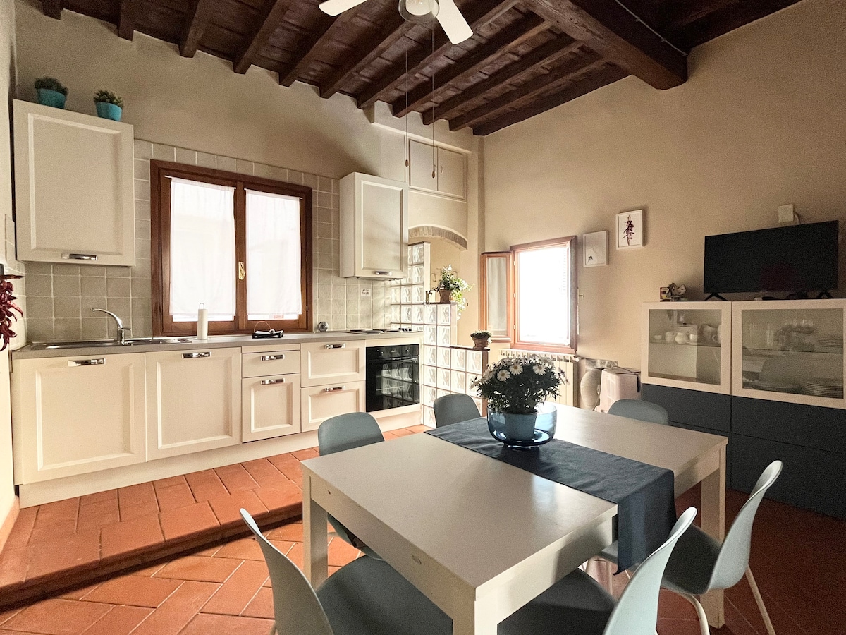 A well-appointed kitchen is presented with light-colored cabinetry and a spacious dining table surrounded by six chairs. Two windows allow natural light to brighten the space, with a decorative bowl of flowers placed at the center of the table.
