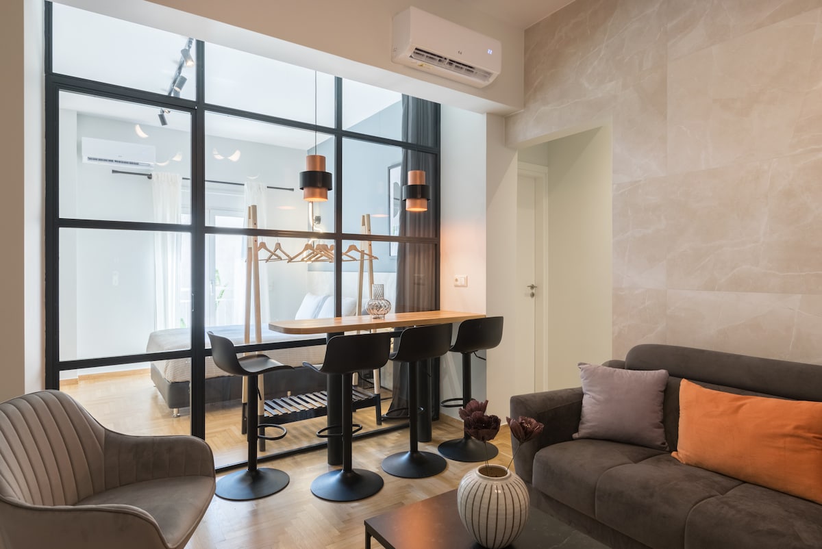 A modern living area features a light wooden dining table surrounded by four sleek black chairs. The space is visually divided by a metal-glass partition, allowing glimpses into additional areas. Soft lighting and neutral tones create a welcoming ambiance, complemented by comfortable seating.