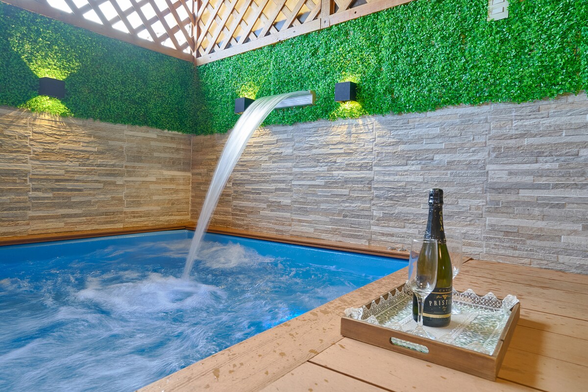 A private jacuzzi is set against a backdrop of textured stone walls and lush green accents. Water flows gently from a wall spout into the inviting blue water. A tray with a bottle of champagne rests on a wooden surface nearby, enhancing the serene atmosphere.