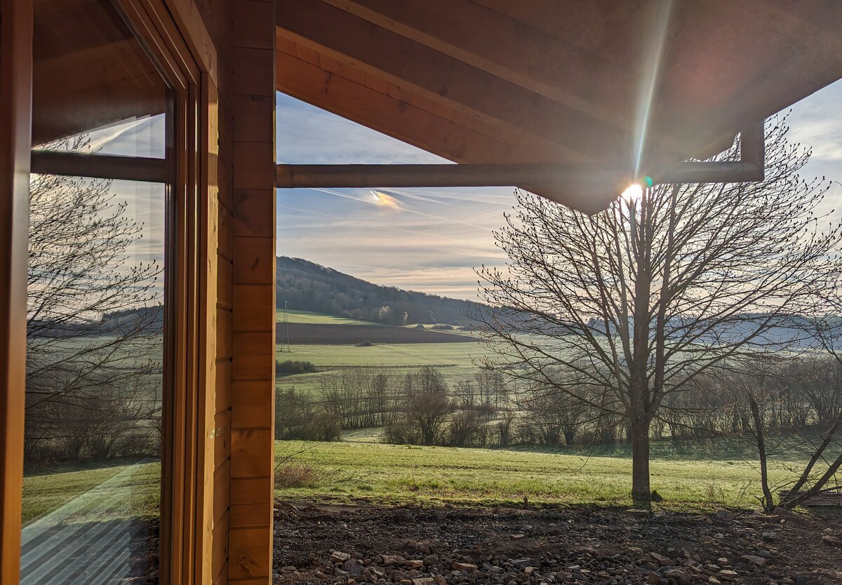 A view from the spacious terrace showcases lush green fields stretching towards distant hills. Sunlight filters gently through the trees, creating a serene atmosphere. Large windows emphasize the connection to nature, inviting the outdoors into the space.