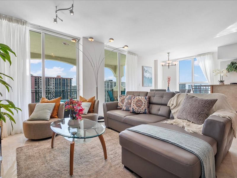 A spacious living area features large glass doors that open to a balcony, allowing natural light to fill the room. A modern sectional sofa and chic armchair provide comfortable seating. A round glass table is centered, accompanied by decorative cushions and a plant for a touch of greenery.