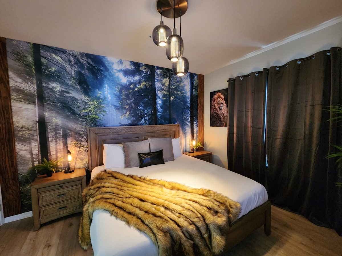 A comfortable bedroom features a spacious bed draped with a soft fur throw. The design is complemented by forest-themed wallpaper, adding a serene touch. Beside the bed, two bedside tables hold decorative lamps, while dark curtains frame the window, enhancing the cozy atmosphere.