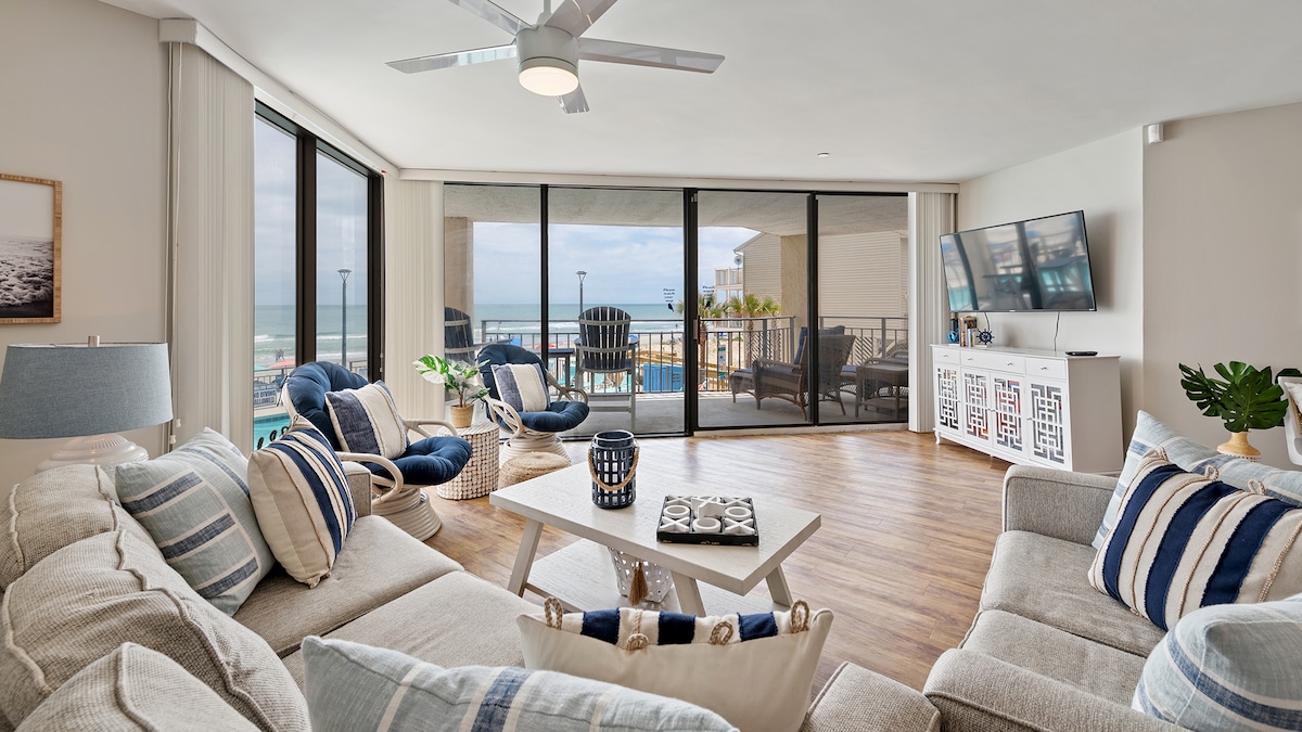 A spacious living area is presented with light wood flooring, featuring a comfortable sectional sofa adorned with blue and white stripes. Large glass sliding doors open to a balcony with pool and ocean views, while a ceiling fan and modern TV complete the bright, airy setting.