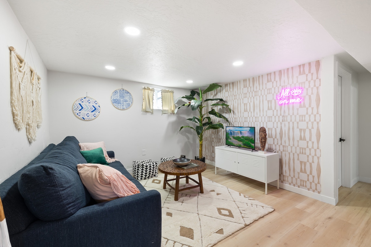A comfortable living area is presented, featuring a dark blue couch with colorful pillows. A round coffee table sits center stage, surrounded by a light rug. A 32" smart TV is positioned on a white cabinet against a patterned wall, complemented by decorative wall hangings and a small indoor plant.