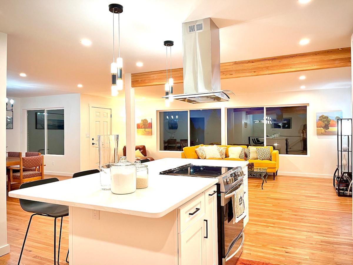 An open-concept kitchen and living area features a modern island with a gas stove, complemented by sleek bar chairs. A vibrant yellow sofa is positioned by large windows, allowing ample natural light. Wood accents embellish the ceilings, enhancing the contemporary design.