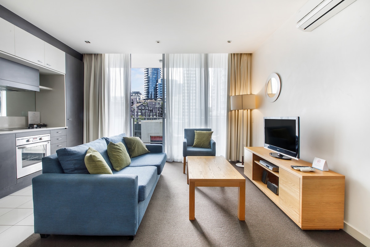 The living area features a comfortable blue sofa and two green armchairs, positioned around a wooden coffee table. Large windows offer natural light and city views, while modern decor adds a stylish touch. An entertainment unit with a television is also present.