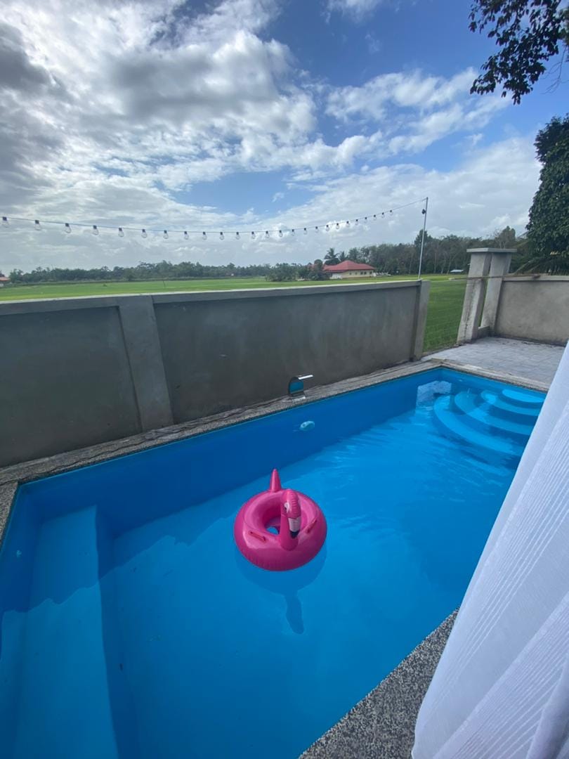 A private pool is visible, featuring bright blue water and a pink inflatable float. Surrounding greenery and open fields can be seen in the background, with decorative lights strung above the pool area, creating a serene and inviting outdoor space.