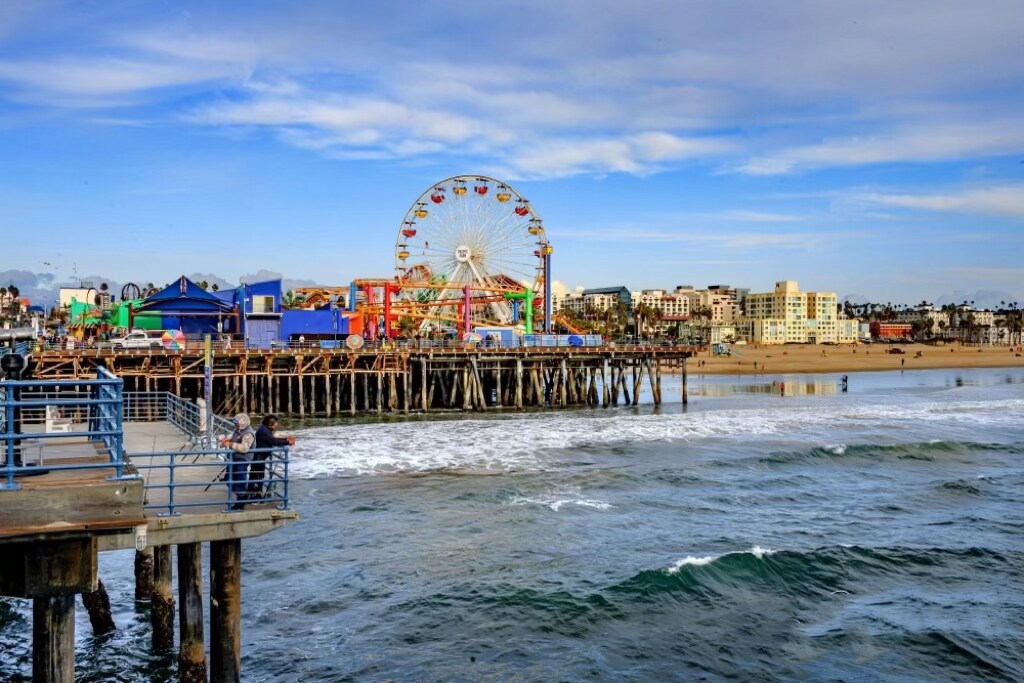 The Santa Monica Pier is captured with its colorful amusement rides overlooking the waves of the Pacific Ocean. The vibrant Ferris wheel stands out against a clear blue sky, while sandy beaches are visible in the background, attracting visitors to the seaside experience.