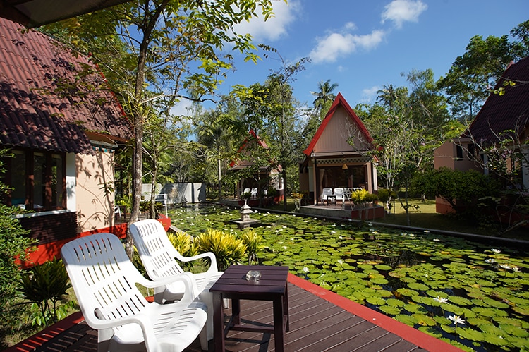 Two white lounge chairs are positioned on a wooden deck, offering a view of a serene garden featuring a pond filled with lily pads. Colorful huts are visible in the background, surrounded by lush greenery and blue skies.
