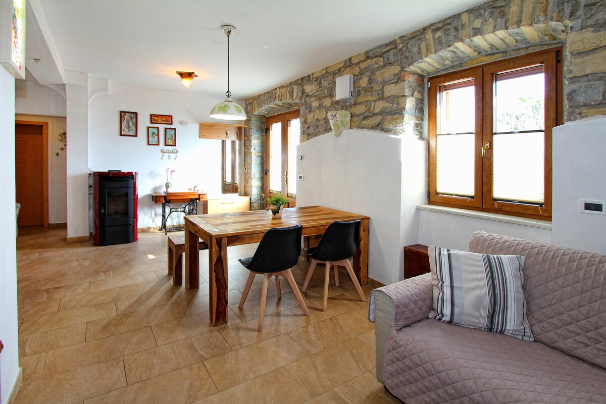 A bright and open living area features a rustic wooden dining table surrounded by stylish black chairs. Natural light enters through the two large windows, highlighting the stone walls and tiled floor. A cozy sofa with a striped cushion is positioned near the wall.