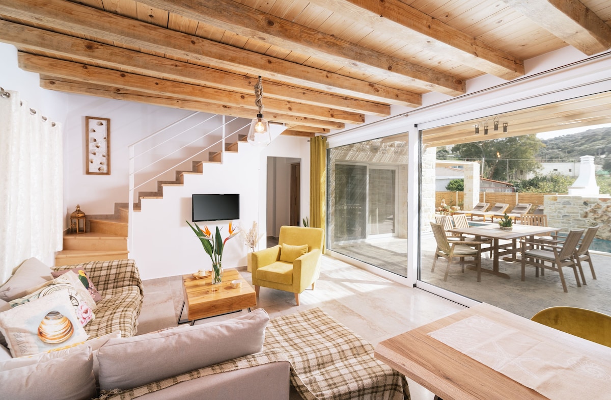 A spacious living area features wooden beams overhead, a comfortable sofa adorned with decorative pillows, and a coffee table topped with a vase of fresh flowers. Large glass doors provide a view of an outdoor dining space, enhancing the flow between indoor and outdoor living.