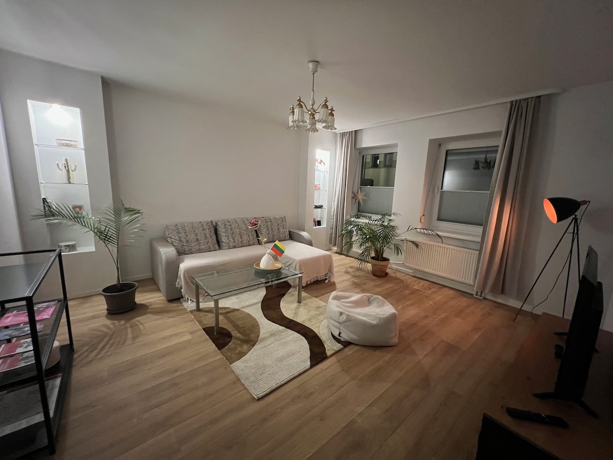 A spacious living area features a neutral color palette, with a comfortable sofa and a glass coffee table atop a decorative rug. Potted plants add a touch of greenery, while light enters through two large windows draped with soft curtains. A stylish floor lamp and a media unit complete the space.
