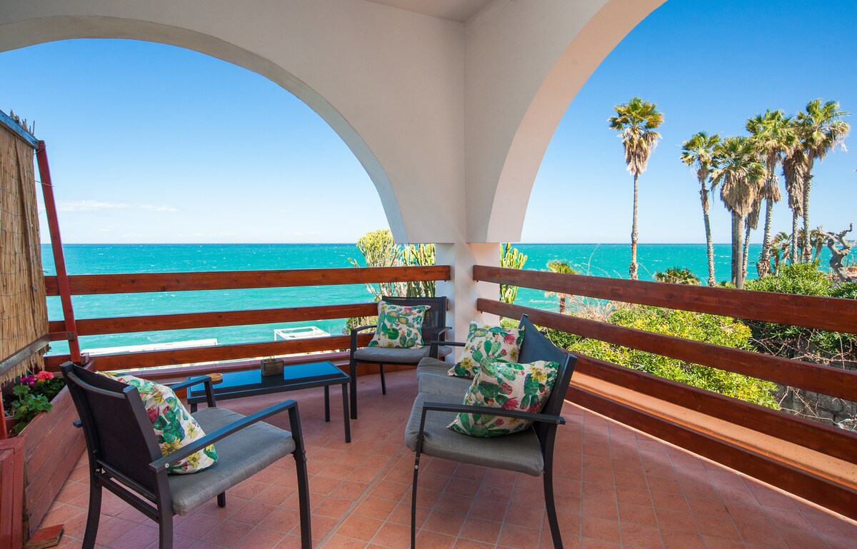 A private terrace offers a view of the turquoise sea and swaying palm trees. Comfortable seating is arranged under an archway, inviting relaxation in a serene outdoor space with sunlight streaming through. Vibrant cushions enhance the seating area, contributing to a coastal setting.